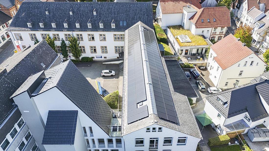 Luftaufnahme eines modernen Stadtviertels mit weißen Gebäuden, dunklen Dächern und Sonnenkollektoren auf dem Dach eines zentralen Gebäudes. Im Innenhof sind mehrere Autos geparkt, und das Gebiet ist von Grün umgeben.